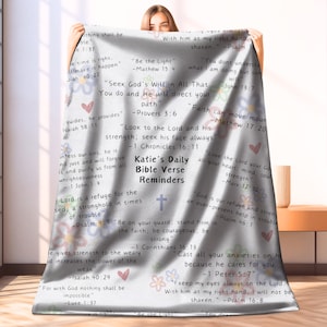 May include: A white throw blanket with a variety of bible verses printed on it. The blanket features a cross, hearts, and flowers in a variety of colours. The text includes verses such as "Be the Light", "Seek God's Will in All That You Do", "Faith Can Move Mountains", and "God is Our Refuge and Strength".