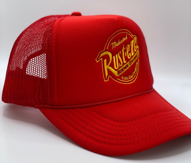 May include: A bright red trucker hat featuring a mesh back and a solid front panel. The front displays an embroidered yellow and orange circular logo with the text Rust-eze, Medicated Bumper Ointment, and Rear-End Formula in a stylized font.