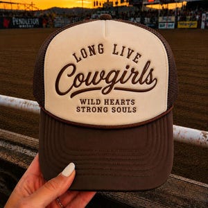 May include: A brown and cream trucker hat featuring a foam front panel with the embroidered text LONG LIVE Cowgirls WILD HEARTS STRONG SOULS in dark brown. The hat includes a solid brown brim and a mesh back panel for ventilation and structure.