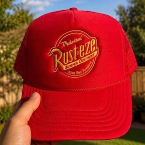 May include: A bright red trucker hat featuring a circular embroidered patch on the front. The patch displays the yellow and red text Rust-eze Medicated Bumper Ointment Rear End Formula. The cap has a solid front panel and a mesh back panel.