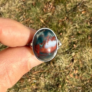 May include: Oval-shaped gemstone ring with a silver band. The gemstone features a mix of red, green, and white colors. The ring is held up against a background of dry grass.