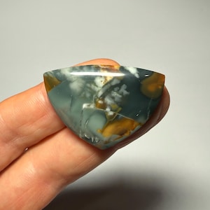 May include: A polished gemstone with a triangular shape. The stone features a mix of colors, including teal, white, and golden-brown, with a marbled pattern. The gemstone is held in a hand, showcasing its unique color variations and natural beauty.