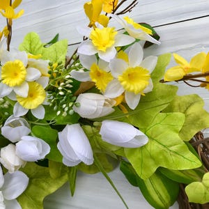 May include: A spring wreath with white and yellow flowers, green leaves, and brown twigs. The wreath is made of natural materials and is perfect for decorating your home for the spring season.