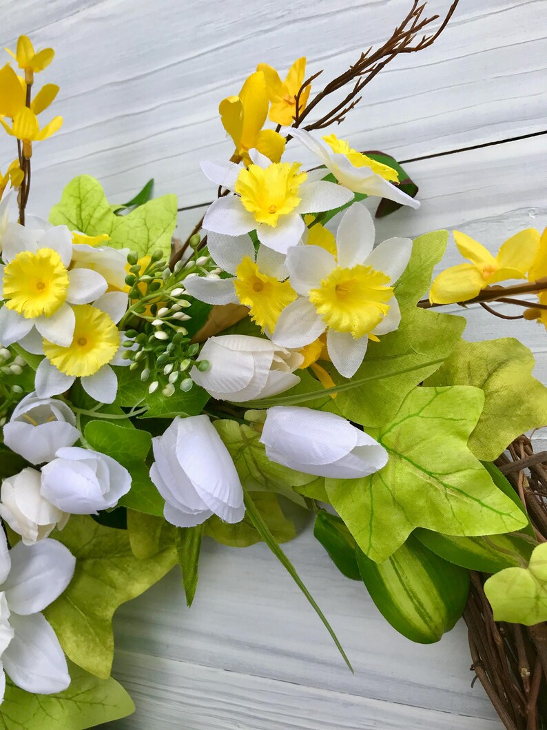 May include: A spring wreath with white and yellow flowers, green leaves, and brown twigs. The wreath is made of natural materials and is perfect for decorating your home for the spring season.