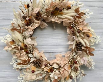 May include: A circular autumn wreath featuring a variety of dried leaves, wheat, pinecones, and berries in shades of cream, brown, and orange. The wreath is arranged on a light gray wooden background, creating a rustic and seasonal decorative piece.