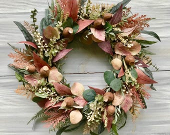 May include: A decorative wreath featuring a circular design with a variety of faux foliage. The wreath includes green, red, and tan leaves, along with small acorns and sprigs of white flowers. The wreath is displayed against a light gray wooden background.