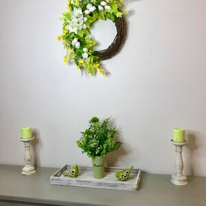 May include: A green wreath with white flowers and yellow accents hangs on a white wall. A green vase with white text that says "Flowers" holds a bouquet of green flowers. Two white candle holders with green candles are on a gray table with a wooden tray with two green birds.