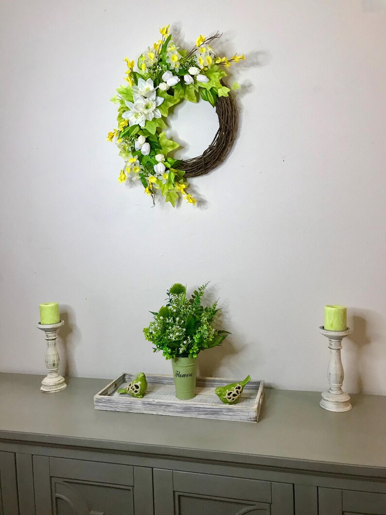May include: A green wreath with white flowers and yellow accents hangs on a white wall. A green vase with white text that says "Flowers" holds a bouquet of green flowers. Two white candle holders with green candles are on a gray table with a wooden tray with two green birds.