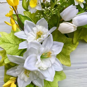 May include: A spring wreath with white daffodils, yellow forsythia, and white tulips. The wreath is made with green leaves and vines.