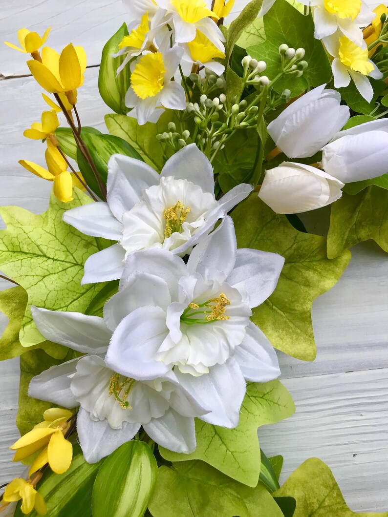 May include: A spring wreath with white daffodils, yellow forsythia, and white tulips. The wreath is made with green leaves and vines.