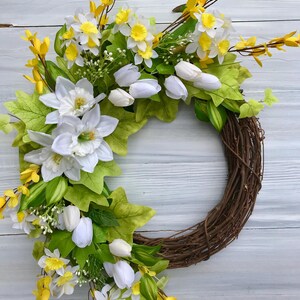 May include: A spring wreath with white tulips, yellow daffodils, and green leaves on a brown vine base.