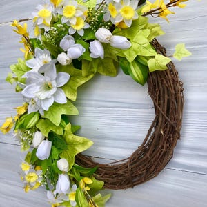 May include: A spring wreath with white and yellow flowers, green leaves, and brown vines. The wreath is made of natural materials and is perfect for decorating your home for the spring season.