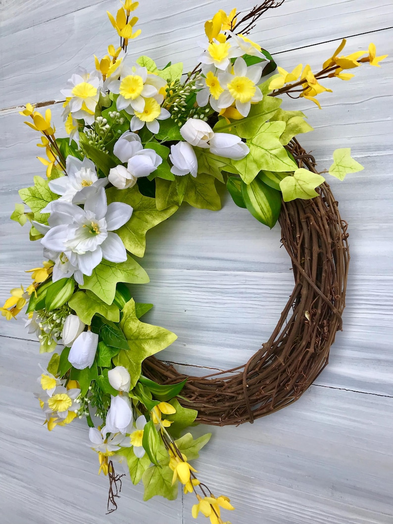 May include: A spring wreath with white and yellow flowers, green leaves, and brown vines. The wreath is made of natural materials and is perfect for decorating your home for the spring season.