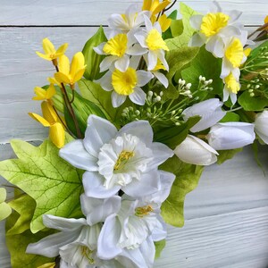 May include: A spring wreath with white daffodils, yellow forsythia, and green leaves. The wreath is made on a brown base.