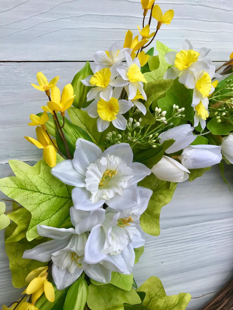May include: A spring wreath with white daffodils, yellow forsythia, and green leaves. The wreath is made on a brown base.