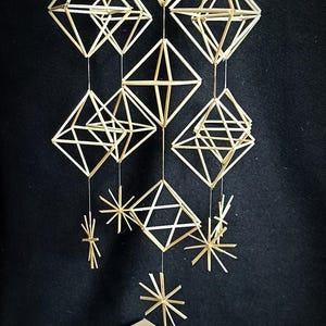 May include: A handmade straw mobile with geometric shapes and starburst ornaments. The natural-colored straw is suspended against a black background. A box with a sunburst design and bundles of straw are in the foreground.