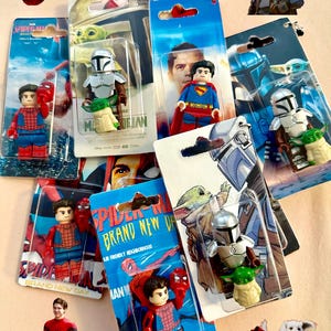 Op de afbeelding: Een verzameling actiefiguurtjes met personages uit Spider-Man, Superman en The Mandalorian. De figuren zitten in hun originele verpakking, met levendige kleuren en gedetailleerde ontwerpen. De afbeelding bevat een vliegende Superman en een hond met een cape.