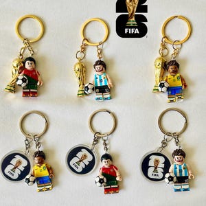 May include: Six keychains featuring miniature soccer player figures in various national team jerseys, each holding a small soccer ball. Some keychains include a gold-colored trophy charm, while others feature a circular tag with the FIFA 26 logo.