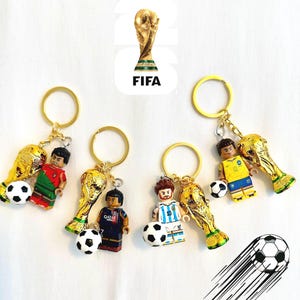 May include: Keychains featuring miniature soccer player figures, a golden trophy, and a soccer ball. Each keychain has a gold-colored ring. The FIFA logo is visible in the background. The figures are wearing different team uniforms.