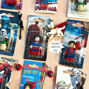 Op de afbeelding: Assortiment van sleutelhangers met superhelden, met personages als Superman, Spider-Man en The Mandalorian. Elke sleutelhanger bevat een kleine bedel en is verpakt op een kleurrijke kaart. De tekst "includes charm for you!" is zichtbaar.