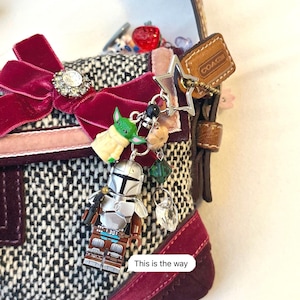 May include: A small, tweed handbag with a burgundy velvet bow and a collection of charms. Charms include a Lego Mandalorian figure, a Baby Yoda figure, a star, and a Coach tag. The text "This is the way" is visible.