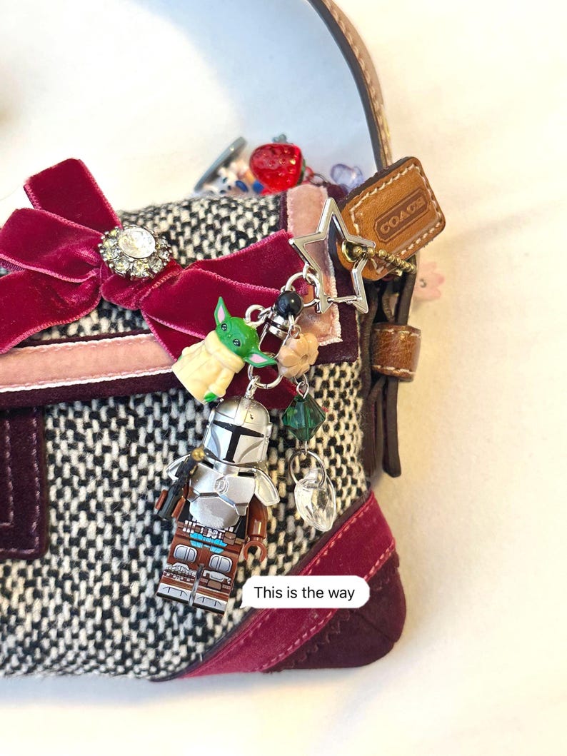 May include: A small, tweed handbag with a burgundy velvet bow and a collection of charms. Charms include a Lego Mandalorian figure, a Baby Yoda figure, a star, and a Coach tag. The text "This is the way" is visible.