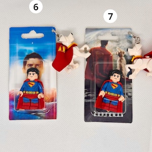 Op de afbeelding: Twee Superman-sleutelhangers. E&eacute;n heeft een Superman Lego-figuur in een blauw pak met een rode cape. De andere sleutelhanger heeft een witte hond met een rode cape. Beide zijn bevestigd aan een blauwzwarte kaart met Superman-afbeeldingen.