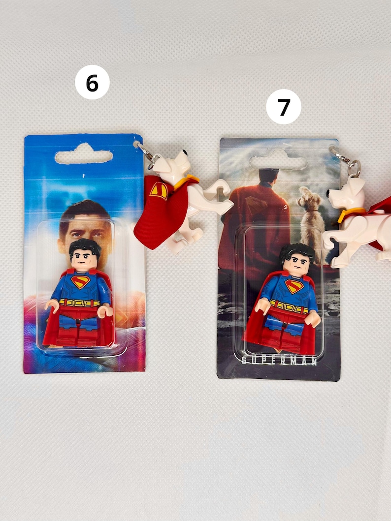 Op de afbeelding: Twee Superman-sleutelhangers. E&eacute;n heeft een Superman Lego-figuur in een blauw pak met een rode cape. De andere sleutelhanger heeft een witte hond met een rode cape. Beide zijn bevestigd aan een blauwzwarte kaart met Superman-afbeeldingen.