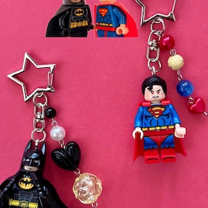 May include: Keychains featuring Batman and Superman Lego figures. The Batman keychain has a black figure with a star-shaped clasp and decorative beads. The Superman keychain has a red and blue figure with a star-shaped clasp and decorative beads.