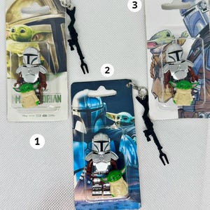 Op de afbeelding: Drie Mandalorian en Grogu sleutelhangers. Elke sleutelhanger heeft een klein figuur van de Mandalorian en Grogu, verpakt op een thematische achtergrondkaart. De Mandalorian heeft zilveren bepantsering en Grogu is groen. Elke sleutelhanger heeft een zwart wapen en een zilveren ketting.