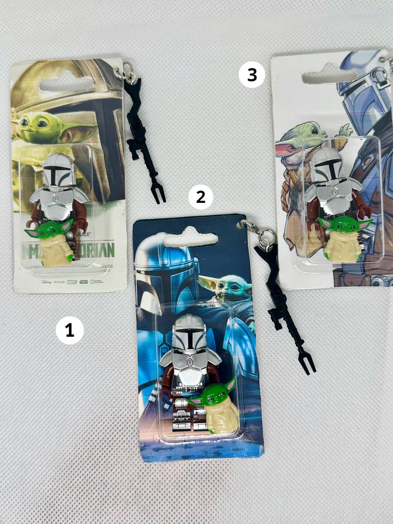 Op de afbeelding: Drie Mandalorian en Grogu sleutelhangers. Elke sleutelhanger heeft een klein figuur van de Mandalorian en Grogu, verpakt op een thematische achtergrondkaart. De Mandalorian heeft zilveren bepantsering en Grogu is groen. Elke sleutelhanger heeft een zwart wapen en een zilveren ketting.