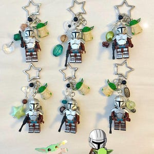 May include: Keychains featuring Mandalorian figures, each with a silver star-shaped top, a small Grogu figure, and decorative beads. The Mandalorian figures are in silver and brown armor, holding a black blaster.