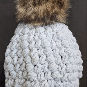 Pode incluir: Um gorro de crochê azul claro com um pompom de pele sintética marrom e felpudo no topo.