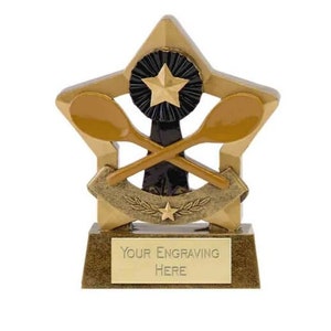 Può includere: Trofeo dorato a forma di stella con due cucchiai di legno incrociati e una stella nera al centro. La base ha una placca dorata con il testo "YOUR ENGRAVING HERE".