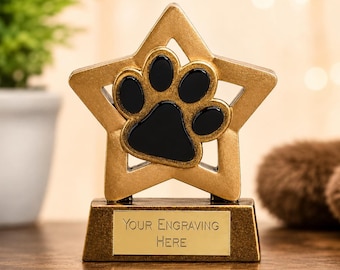 Può includere: Trofeo a forma di stella color oro con un'impronta di zampa nera al centro. Il trofeo ha una base rettangolare con la scritta "Your Engraving Here" su una placca dorata. Lo sfondo è sfocato.