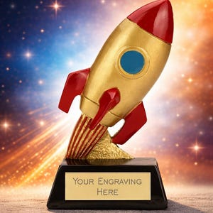 May include: A gold-coloured rocket trophy with red accents, a blue porthole, and a black base. The base has a gold plate that reads "YOUR ENGRAVING HERE". The rocket is angled upwards, suggesting flight.
