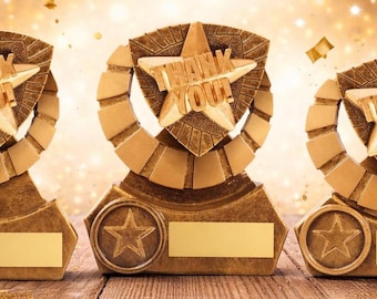 May include: Three gold-colored trophies with a star design and the words "THANK YOU" on the front. Each trophy has a star emblem and a blank rectangular space for personalization. The background features a bokeh effect with gold confetti.