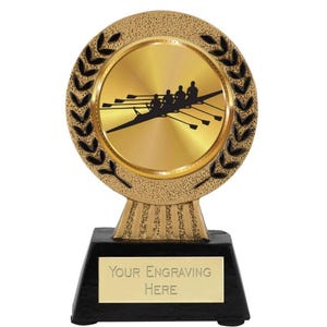 Peut inclure: Trophée de couleur dorée avec une base noire. Le trophée présente un sommet rond de couleur dorée avec une silhouette noire d'une équipe d'aviron. Le sommet est entouré d'une couronne de laurier noire. La base a une plaque dorée avec le texte "YOUR ENGRAVING HERE".