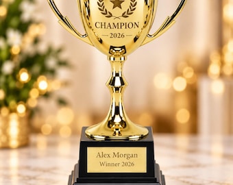 May include: A shiny gold trophy cup with two handles, featuring an engraved star, laurel wreath, and the text CHAMPION 2026. The trophy sits on a tiered black base with a gold plaque reading Alex Morgan Winner 2026 in black lettering.