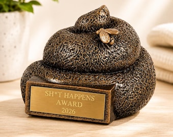 Könnte beinhalten: Eine bronzefarbene dekorative Skulptur in Form eines gewundenen Haufens mit strukturierter Oberfläche und einer kleinen Fliegenfigur auf der Oberseite. Ein rechteckiges goldenes Schild auf der Vorderseite trägt den Text SH*T HAPPENS AWARD 2026.