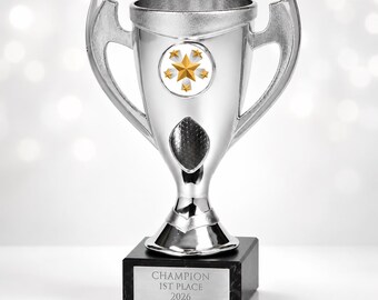 May include: A silver-colored trophy cup with two handles, featuring a circular emblem with five gold stars on the front. The trophy is mounted on a black rectangular base with a metal plate engraved with the text CHAMPION 1ST PLACE 2026.