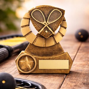May include: A gold squash trophy featuring crossed rackets on a shield, a star, and a blank nameplate. A squash ball and racket are in the foreground. The trophy is on a wooden surface.