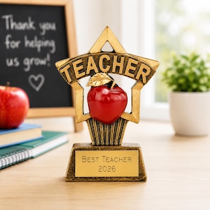 May include: A gold-coloured trophy featuring a star-shaped frame with the word TEACHER embossed across the top. A glossy red apple with a gold leaf sits in the centre. The base has a plaque with the text BEST TEACHER 2026 engraved in black lettering.