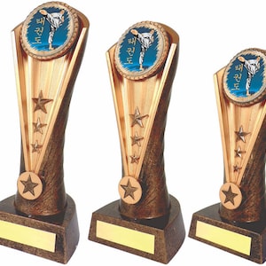 Puede incluir: Tres trofeos de color dorado con un logotipo de Taekwondo azul y blanco en la parte superior. Cada trofeo tiene una estrella dorada en la base y tres estrellas doradas en el lateral.