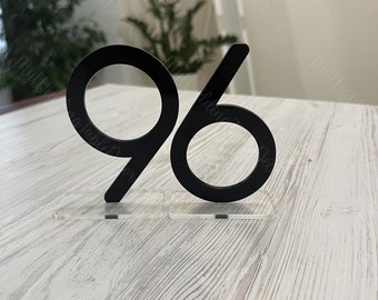 May include: A black acrylic table number, displaying the number 96, on a clear base. The font is bold and sans-serif. The background features a light wooden surface and blurred greenery.
