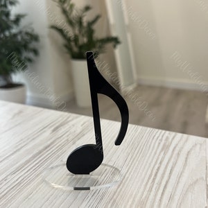 May include: Black music note table decor on a clear acrylic base. The note is standing on a light wood surface.