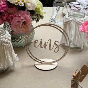 Op de afbeelding: Een houten tafelnummer met het woord "eins" in een dubbele cirkel, geplaatst op een ronde basis. De tafel is gedekt met bloemstukken in glazen vazen, met macramé bedekte potten en glaswerk. De bloemen zijn roze, wit en groen.