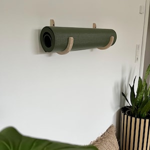 May include: An olive green yoga mat, rolled up and held by a wooden wall mount. The mount has two curved wooden supports. The mat is mounted on a white wall. A potted plant is in the background.