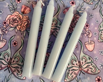 May include: Four pale blue taper candles with white wicks are arranged diagonally. The candles are on a lavender fabric with a repeating pattern of butterflies, roses, and other floral elements. The candles are a light, pastel color.