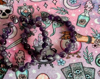 May include: A beaded bracelet made of purple amethyst stones. The bracelet is displayed on a pink and purple patterned surface with occult-themed illustrations, including cats, mushrooms, and tarot cards.